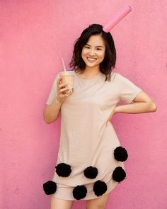 a woman in a pom - pom dress holding a drink and standing against a pink wall