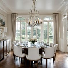 a dining room table with white chairs and chandelier in front of large windows