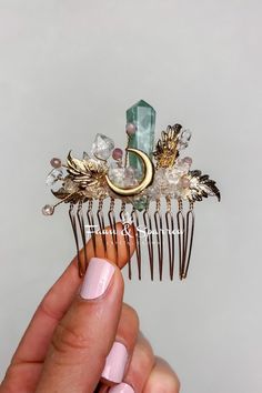 a hand holding a hair comb with crystals and leaves on it's side, in front of a gray background