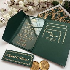 an open green and gold wedding card with two coins next to it on a table