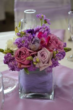 a square vase filled with purple and white flowers on top of a pink table cloth