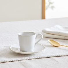 a white cup and saucer sitting on top of a bed next to two gold spoons