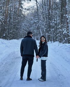 a man and woman holding hands walking down a snow covered road in the woods on a cold winter day