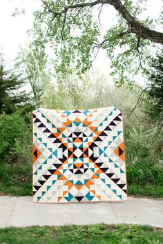 an orange, white and blue quilt sitting on top of a cement sidewalk next to trees