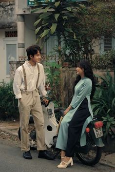 a man standing next to a woman sitting on a scooter in the street