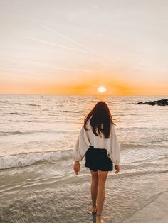 a woman walking on the beach at sunset with her back to the camera, looking out into the water