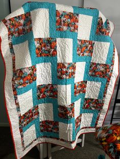 a large blue and white quilt on a metal stand in front of a chair with an orange flowered pillow