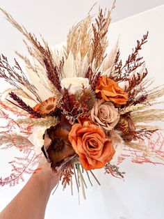 a hand holding a bouquet of flowers on top of a white surface with dry grass