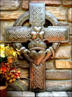 a metal cross on the side of a stone wall with flowers in front of it