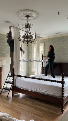 two people standing on top of a bed in front of a chandelier and window