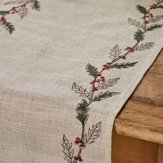 an embroidered table runner with holly and red berries on the edge is sitting on a wooden bench