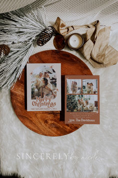 two christmas cards sitting on top of a wooden plate next to a candle and pine cone