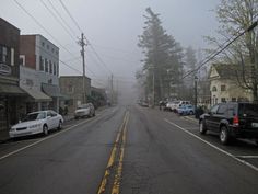 cars are parked on the street in front of buildings and trees, with fog covering the sky