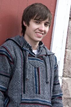 a young man standing in front of a red wall wearing a gray and blue sweater