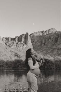 a pregnant woman standing in front of a body of water with mountains in the background