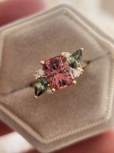 a pink ring with green and white stones on it's side in a box