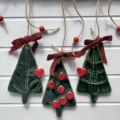 three ceramic christmas trees hanging from strings on a white wall with red berries and twine