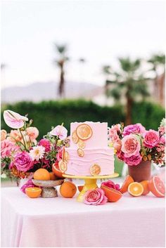 a pink cake sitting on top of a table covered in flowers and fruit next to potted plants