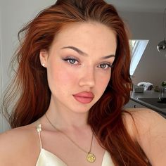 a woman with long red hair wearing a white top and gold necklace, posing for the camera