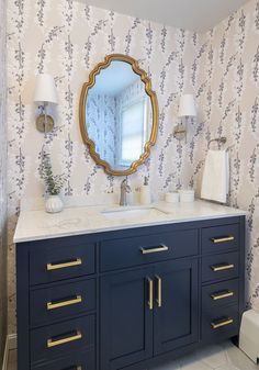 a bathroom vanity with blue cabinets and gold trim on the mirror above it is decorated in floral wallpaper