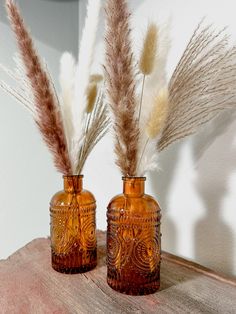 two brown vases sitting on top of a wooden table next to each other with dried grass in them