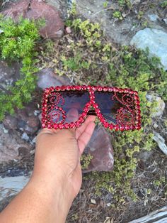 a hand is holding up a pair of sunglasses with red beads on it and some rocks in the background