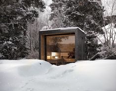 a small wooden cabin in the middle of snow covered trees and bushes, with a person sitting inside