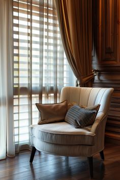 a white chair sitting in front of a window covered with curtains and pillows on top of a hard wood floor