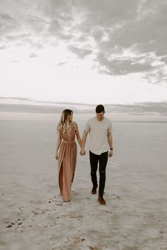 a man and woman holding hands while walking on the beach in front of an overcast sky