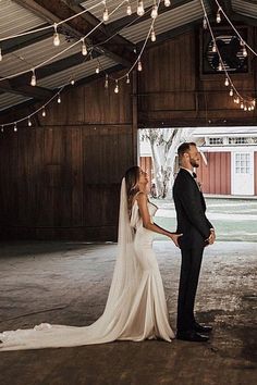 a bride and groom standing in an old barn looking at each other with lights hanging from the ceiling