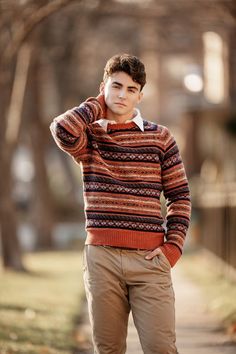 a young man standing on a sidewalk wearing a sweater and khaki pants with his hands in his ears