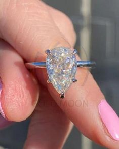 a woman's hand holding a ring with a pear shaped diamond on it and pink nail polish