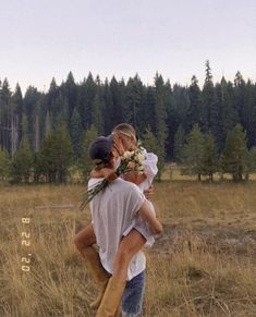 a man carrying a woman in the middle of a field with pine trees behind him