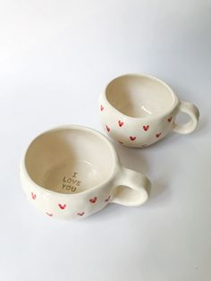 two white cups with red hearts on them sitting side by side, one has the words i love you written on it