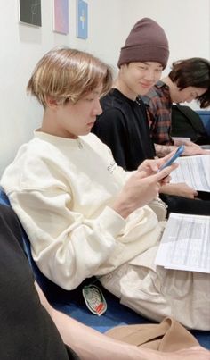 three young men sitting in a room looking at papers on their cell phones while one man is holding a pen