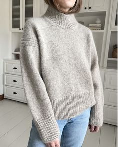 a woman standing in front of a white cabinet wearing a gray sweater and blue jeans