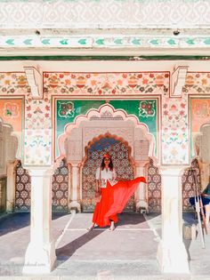 a woman standing in front of a colorful building with arches and pillars, wearing a long red skirt