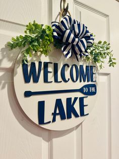 a welcome to the lake sign hanging on a white door with green leaves and a blue bow