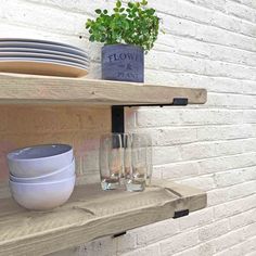 a shelf with glasses and plates on it next to a brick wall in front of a potted plant