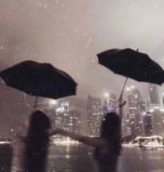 two people holding umbrellas in front of a cityscape at night with rain falling on them