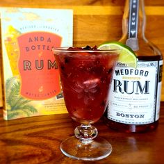 a glass filled with red liquid next to a bottle of rum and a book on a wooden table