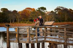 two people standing on a dock near the water with a camera in their hand and another person taking pictures