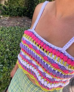 a woman wearing a colorful crochet tank top and green plaid shorts standing in front of bushes