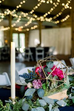 an arrangement of flowers and greenery sits on a table in front of a candle