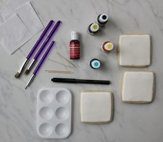 the supplies needed to decorate cookies are laid out on a marble counter top, including paint and brushes