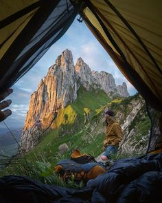 a man sitting in a tent on top of a lush green hillside next to a tall mountain