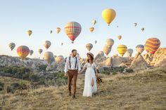 a man and woman are walking through the field with hot air balloons in the sky