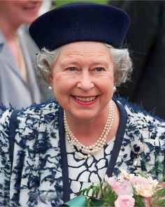 an older woman wearing a blue hat and dress with flowers in her lap smiling at the camera