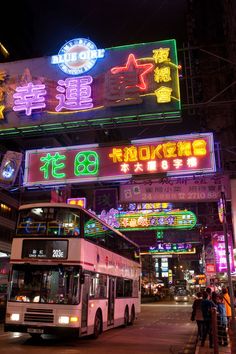 a double decker bus driving down a city street at night with neon signs above it