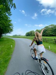 a woman riding a bike down a road next to a lush green field with trees